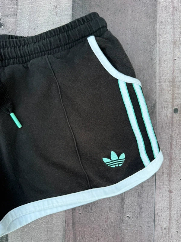 Black Adidas track shorts with mint green stripes and logo, laid flat on a gray wooden plank background.