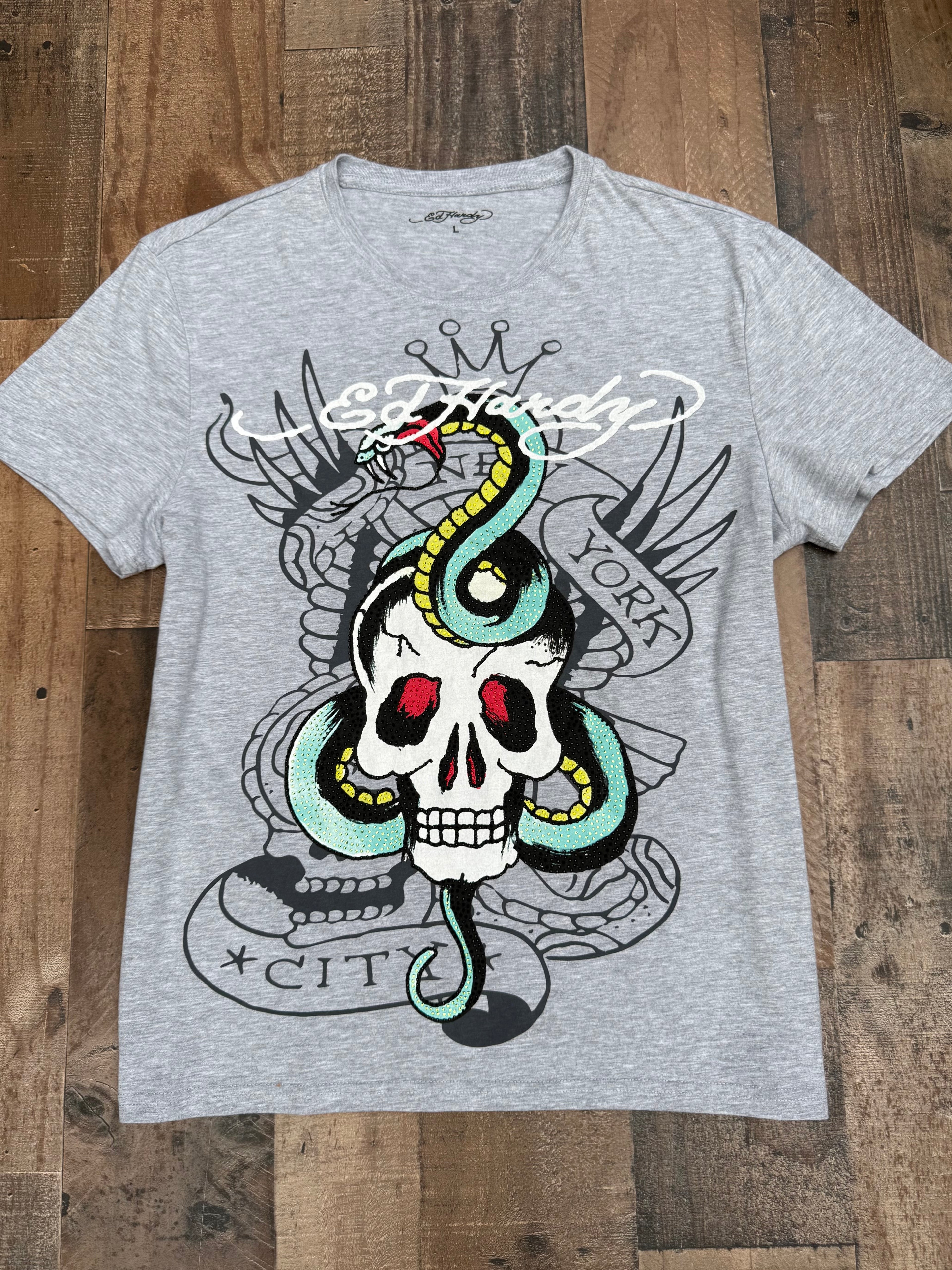 A heather gray Ed Hardy graphic t-shirt featuring a detailed tattoo-style design of a skull with a green and yellow snake, red eyes, and the Ed Hardy brand name across the chest, laid flat on a wooden floor.