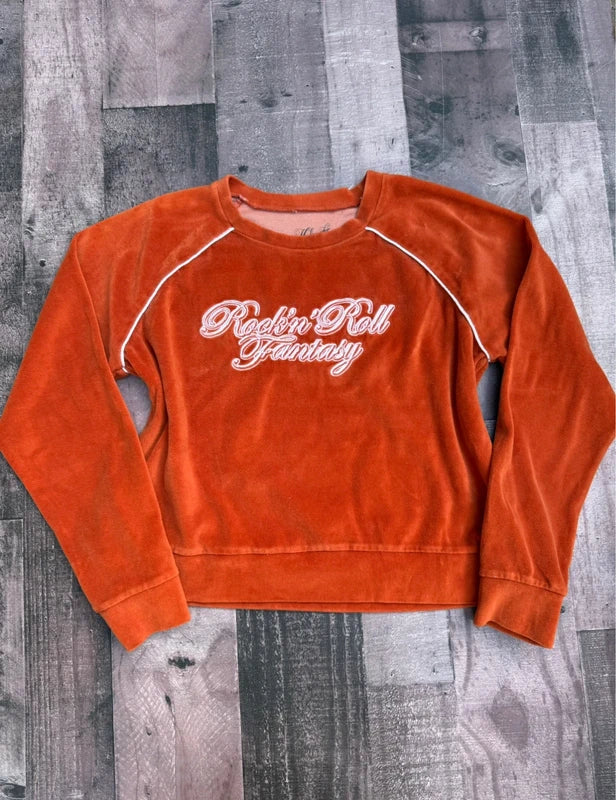 An orange velvet long-sleeve sweatshirt with white embroidered text reading 'Rockin' Roll Fantasy' across the chest, laid flat on a gray wood plank background.